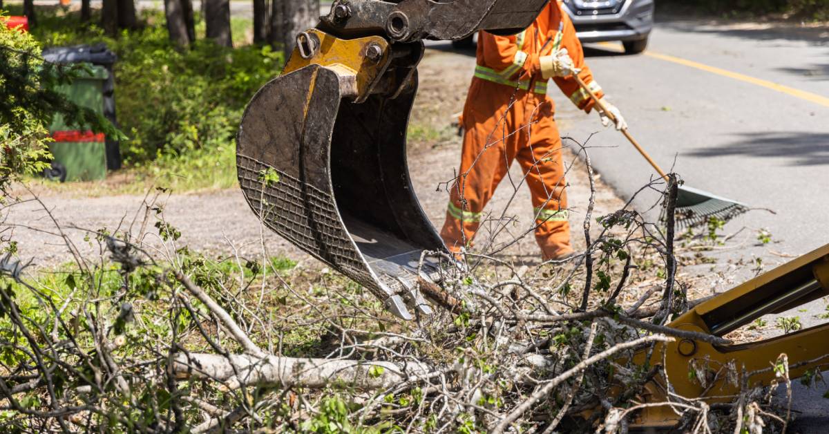 A person in protective gear rakes a driveway. The arm of an excavator cleans up branches from a yard.